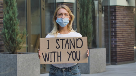 Caucasian female protester activist wearing a medical mask holds a cardboard poster with the slogan I STAND WITH YOU. A protest against racism and violence during quarantine. Stop racism concept.の写真素材
