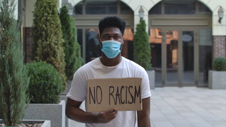 Young African American man in a medical mask stands with a cardboard poster NO RACISM in a public outdoor place. An anti racist movement to protest against injustice. Street demonstration.の写真素材