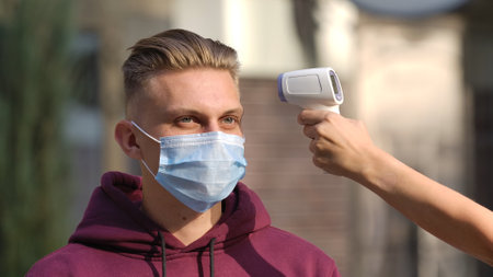 Portrait of young man in medical mask whose temperature is measured with contact less thermometer.