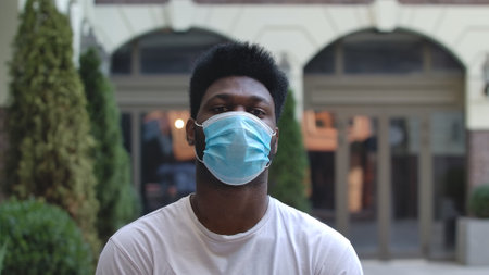 Portrait of young African American man in medical mask looking at the camera. Stylish brunet in a white T shirt on the background of the street outdoors.の写真素材