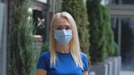 Portrait of young woman in protective medical mask looking into the camera. Blonde in blue blouse on...