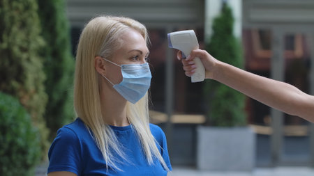 Portrait of young woman in medical mask whose temperature is measured with contactless thermometer.の写真素材