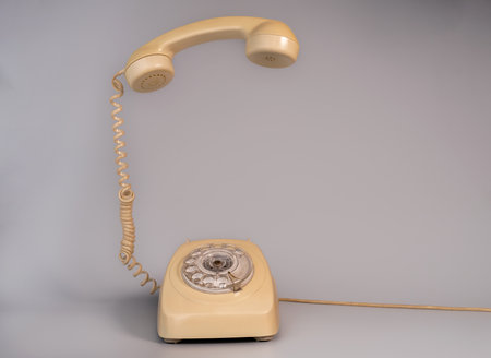 Old beige aged rotary telephone with removed hanging receiver on grey background. Vintage landline home phone with dial, twisted cable and reciever handset. Photo in retro style.の写真素材