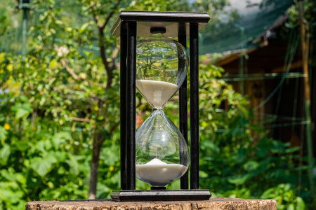 Old hourglass on wooden deck on a blurred background of a green garden. Glass sandglass in black...