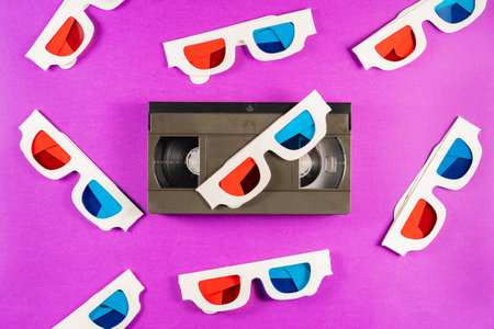 Old white paper 3d glasses with blue red lenses and black videotape vhs on purple background. Retro...
