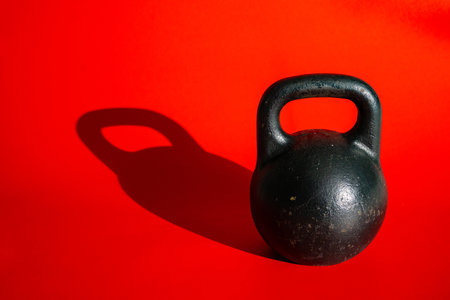 Round black kettlebell with handle on bright red background with shadow. Old heavy metal weight kettlebell for sports, bodybuilding, weightlifting, crossfit. Fitness, power lifting, strength training.の写真素材