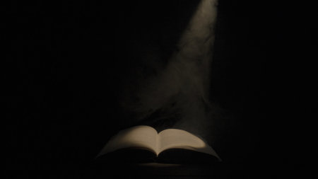 An open book illuminated by a beam of light and smoke on a black isolated studio background. An old textbook with a spot of light on the pages in the darkness of the library. Mysterious open book.の写真素材