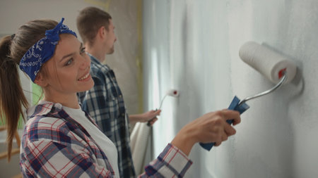 Young couple painting wall with white paint using paint rollers. Pretty woman and man in checkered shirts enjoying renovations in apartment and smile. Concept of repair, finishing work.の写真素材