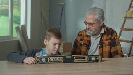 Grandpa is showing and teaching his grandson how the water level works. A teenager with interest examines the level lying on the table. The concept of repair in the apartment, learning.の写真素材