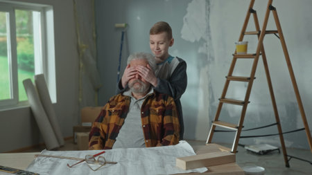 Grandpa is planning repairs sitting at the table. Grandson sneaks up behind and closes his grandfathers eyes with his hands. The concept of family, joint renovation in the apartment.の写真素材