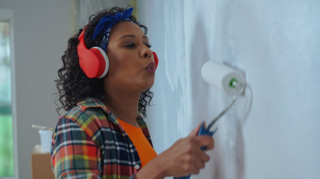 African American woman in red headphones paints wall with white paint using paint roller and enjoy the music. Black female making repairs in an apartment. Close up.の写真素材