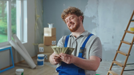 Repairman worker in blue overalls is counting money and smiling. Portrait of a redhead man is posing...