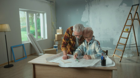 Elderly man and woman are looking through sheet with plan of an apartment and discussing renovation project. Happy aged couple is sitting at a table and planning the improvement of their home.の写真素材