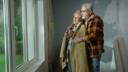 An elderly man hugs an aged woman wrapped in a plaid and with a cup in her hands. A couple of pensioners are dreaming about something looking out the window and smiling happily. Cozy home environment.の写真素材