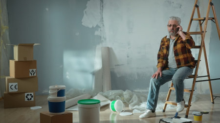 Tired elderly man is sitting on a ladder and calling a master to repair an apartment using a mobile phone. Aged pensioner solved problem with help of the master. Concept of repair.の写真素材