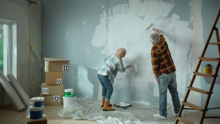 Elderly man and woman are painting wall with white paint using paint rollers. Couple of pensioners are making repairs to their apartment, in the background of window, stepladder, cardboard boxes, wallpaper and cans of paint. Back view.の写真素材