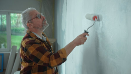 Elderly man is painting wall with white paint using paint roller. Male pensioner is making repairs to his apartment against the backdrop of a window with bright sunlight. Side view.の写真素材