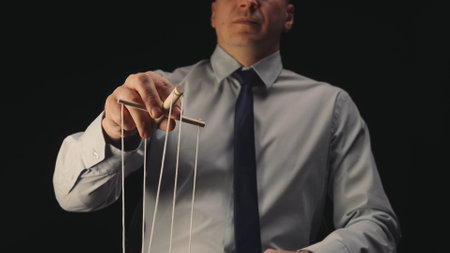 A businessman in a gray shirt and black tie controls a puppet with a wooden manipulator and strings. A puppeteer controls a doll marionette on a black isolated background. Close up.の写真素材