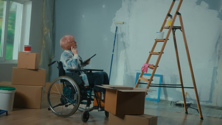 An elderly disabled woman in a wheelchair plans renovation using a digital tablet. Room with window, ladder, cardboard boxes, wallpaper rolls, paint bucket.の写真素材
