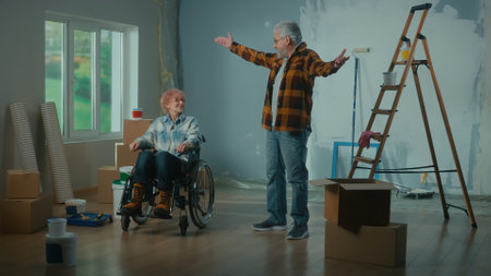 An elderly man and older woman in a wheelchair. An elderly family couple communicate and plans repairs. Room with a window, ladder, cardboard boxes, buckets of paint, rollers, brushesの写真素材