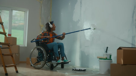An African American woman in white headphones on a wheelchair paints a wall with white paint using a long roller. A young black woman with a disability makes repairs in room and enjoys music.の写真素材