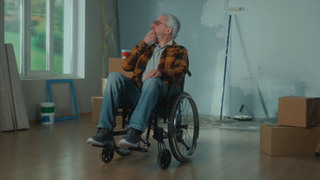 An elderly disabled man in a wheelchair. The pensioner is thinking about doing home repairs. Room with window, cardboard boxes, wallpaper rolls, roller and paint.の写真素材