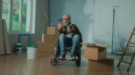 An elderly disabled man in a wheelchair talks on a video call using a mobile phone. Retired person with disabilities. A room with window, wallpaper rolls, cardboard boxes and a bucket of paint.の写真素材