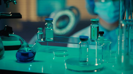 Shelf with glass test tubes, flasks and vials with vaccine and drugs. Medical office in laboratory or hospital with blue light. In the background is a female doctor. Close up.の写真素材