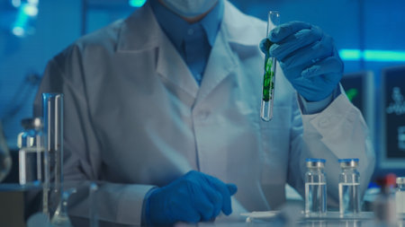 A male doctor in a white coat and blue gloves holds in his hand a glass test tube with a green substance dissolving in a clear liquid. Study of samples, chemical reaction. Close up, blue light.の写真素材