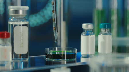 A drop of clear liquid drips from a glass pipette into a petri dish with a green substance. Nearby on the table are glass vials with a vaccine or medicine. Medical background, close up.の写真素材
