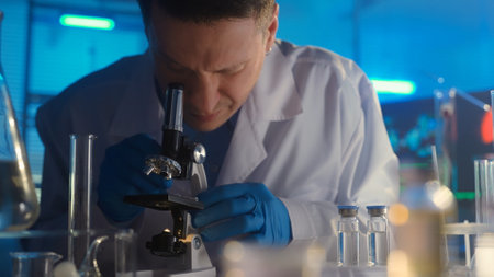 A male researcher in a white gown and blue gloves is examining a sample in a microscope. Laboratory or hospital with many test tubes and glass flasks. In the background is a computer monitor.の写真素材