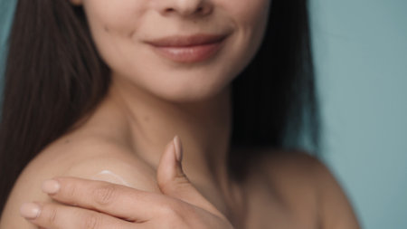 With slow massage movements, the woman applies the cream on her shoulder. A woman takes care of her skin close up on a blurred blue background. The concept of skin and body care. Natural beauty.の写真素材