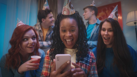 A group of young people in party hats at a home birthday party. Girlfriends, sitting on the couch, are surprised, shocked and joyfully looking at the phone, which is held by an African American girl.の写真素材