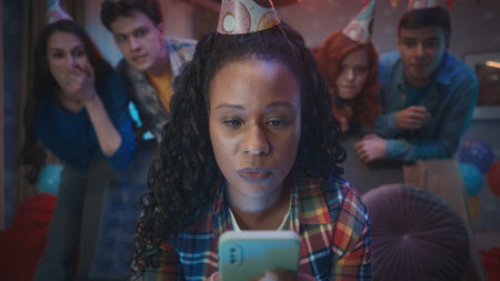An African American woman sits on a couch, looks at her phone, plays, looks at photos, videos. The girls friends stand behind her. A group of people in festive hats at a house party.の写真素材