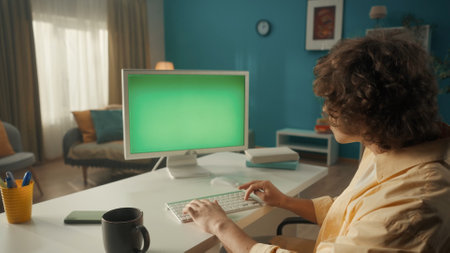 A young curly guy works on a computer with a green screen close up. A man sits at a table with a...