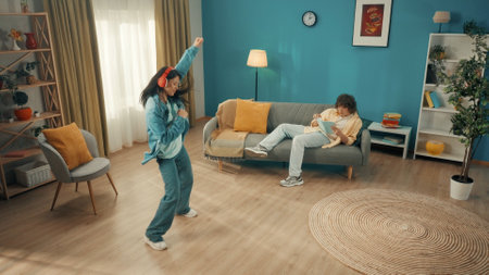 Young curly man is reading a book on the sofa. An Asian woman next to him dances vigorously wearing red wireless headphones. The concept of relax time at home, leisure.の写真素材