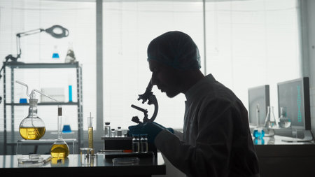 A young scientist in a research laboratory examines a sample on a micropreparation using a microscope. Side view of the dark silhouette of a man sitting in front of a microscope.の写真素材