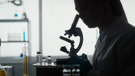Side view of a dark silhouette of a female scientist looking under a microscope, doing an analysis of a test sample close up. Ambitious biotechnology specialist, working with advanced equipment.の写真素材