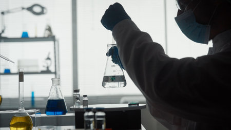 Dark silhouette of a male scientist conducting a scientific experiment close up. A man using a pipette will add a green reagent to a glass flask with a clear liquid.の写真素材