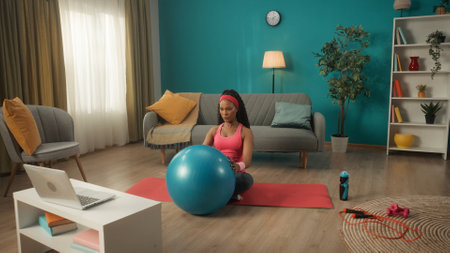 An African American woman in a tracksuit and headband sits crosslegged on a sports mat in the living room. A young woman holds a fitness ball in front of her and looks at a laptop on the table.の写真素材