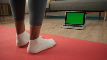 An African American woman is standing on a sports mat in front of a green laptop screen. Female legs in white socks in front of a green laptop screen close up. Advertising area, workspace mock up.の写真素材