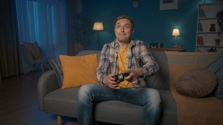 A man plays a consular game with a game controller while sitting on the couch at home in the...