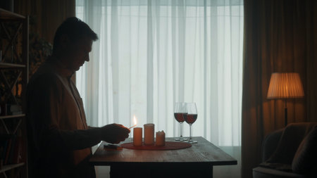 The man is getting ready for a date. The dark silhouette of a man lights candles with matches. Candles and glasses with red wine on the table.の写真素材