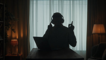 Dark silhouette of a man in wireless headphones at a table in front of a laptop. A man works on a laptop, listens to music, dances.の写真素材