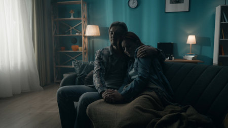 A couple is sitting on a sofa in a dark room. A man holds a woman by the hand, hugs. Support for the couple, joint problem solving, reconciliation and understanding in relationships.の写真素材
