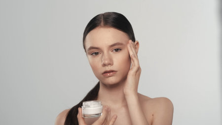 Attractive woman with smooth skin holding a small jar and applies cream to her faceの写真素材