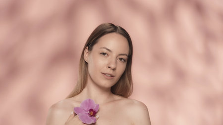 A woman touches an orchid flower to the clean, smooth skin. Portrait of a woman with an orchid flower in her hand in the studio on a pink background close up.の写真素材
