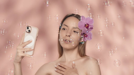 A woman with bare shoulders and orchid flowers in her hair takes a selfie. Seminude woman with a...