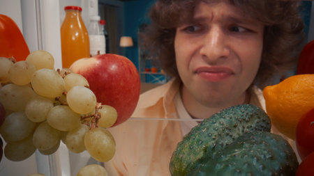 A man stands at the open refrigerator with healthy food. The upset man grimaces, not finding...