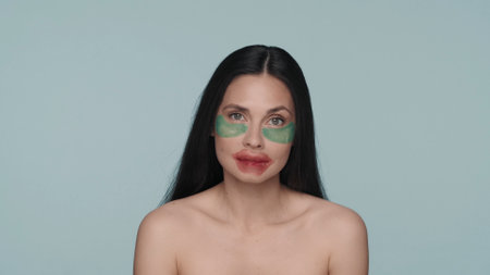 Brunette woman doing facial skin care procedures. Woman with hyaluronic green patches under her eyes and red patches on her lips in the studio on a blue background close up.の写真素材